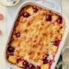 Top-down view of a cherry pineapple dump cake with a golden-brown crust, vibrant cherries, and pineapple pieces baked in a white dish, surrounded by whipped cream and fresh cherries on the table.