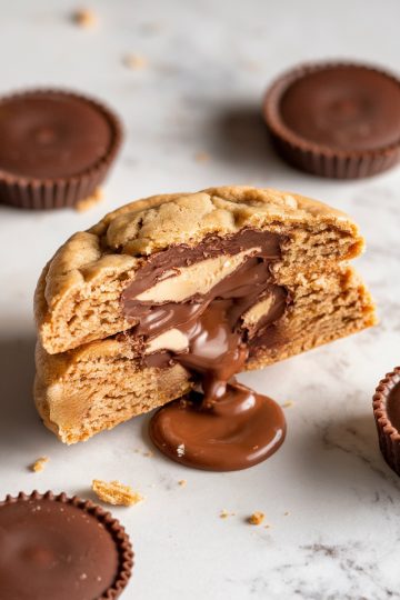 A close-up of a peanut butter chocolate chip cookie split in half, showcasing a gooey, melted chocolate and creamy peanut butter filling that oozes onto a marble countertop. Surrounded by whole peanut butter cups, this indulgent cookie is a treat for chocolate and peanut butter lovers, perfect for dessert inspiration and stuffed cookie recipes.