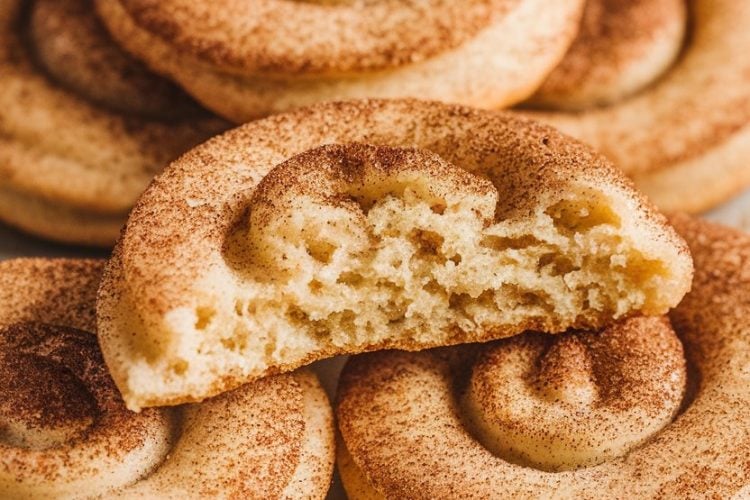 A plate of golden-brown snickerdoodle cookies with a distinctive spiral shape and a crackled, cinnamon-sugar coating. One cookie is broken open, showing a soft, chewy center, perfect for a sweet treat or holiday dessert.