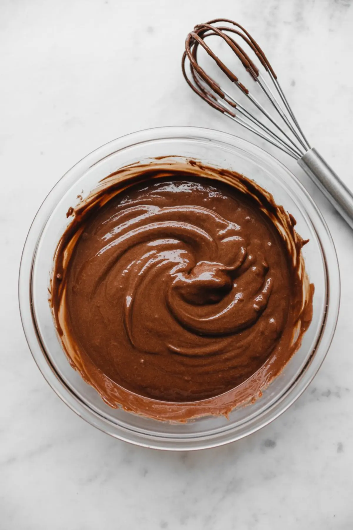 Glass bowl filled with rich chocolate cupcake batter freshly whisked to a smooth consistency, placed on a marble surface with a metal whisk beside it.