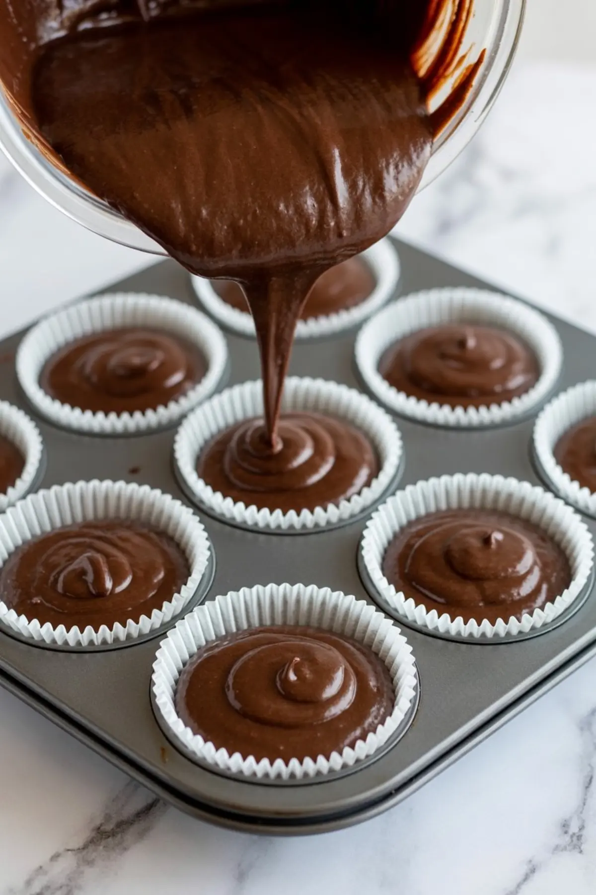 Chocolate cupcake batter being poured from a mixing bowl into paper-lined muffin tin wells, showing a smooth flow of batter for even filling.