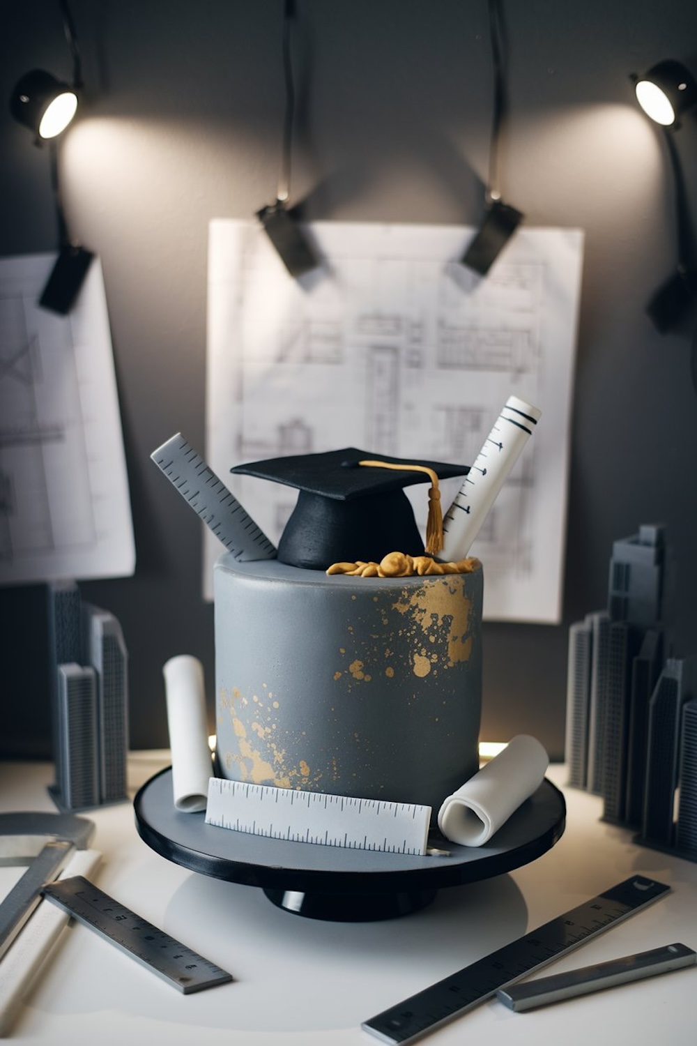  A modern gray cake with gold accents, featuring fondant rulers, blueprints, and architectural tools, topped with a graduation cap, ideal for celebrating an architect or engineer graduate.