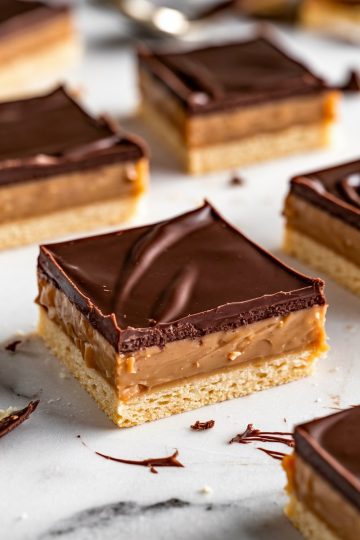 A close-up of a millionaire's shortbread cookie, showcasing its three layers: a buttery shortbread base, a smooth caramel center, and a thick, glossy chocolate top. The square slices are neatly arranged on a marble surface, with bits of chocolate scattered around.