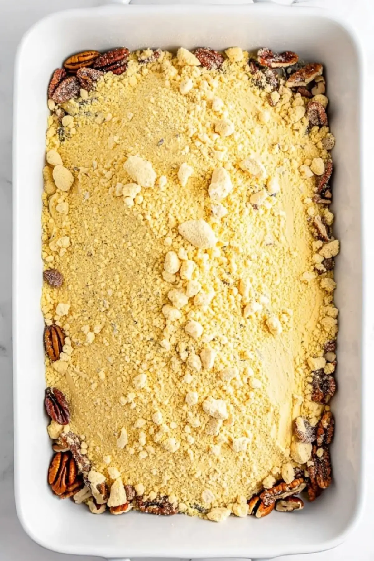 A white baking dish containing a layer of yellow cake mix sprinkled over chopped pecans, capturing a simple dessert preparation scene ideal for easy holiday baking ideas.
