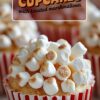 Close-up of a cupcake in a red-and-white striped wrapper, topped with lightly toasted mini marshmallows, resembling popcorn in appearance.