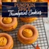 This image shows a combination of two main sections. At the top, it features a close-up of pumpkin spice thumbprint cookies arranged on a cooling rack, highlighting the cinnamon-sugar coating and creamy pumpkin filling swirled in the center. Below, the text "easy pumpkin Thumbprint Cookies recipe" is displayed in vibrant orange and purple fonts over a beige background. At the bottom of the image, a plate piled with more pumpkin thumbprint cookies emphasizes their soft texture and fall-inspired design, with blurred autumn leaves in the background