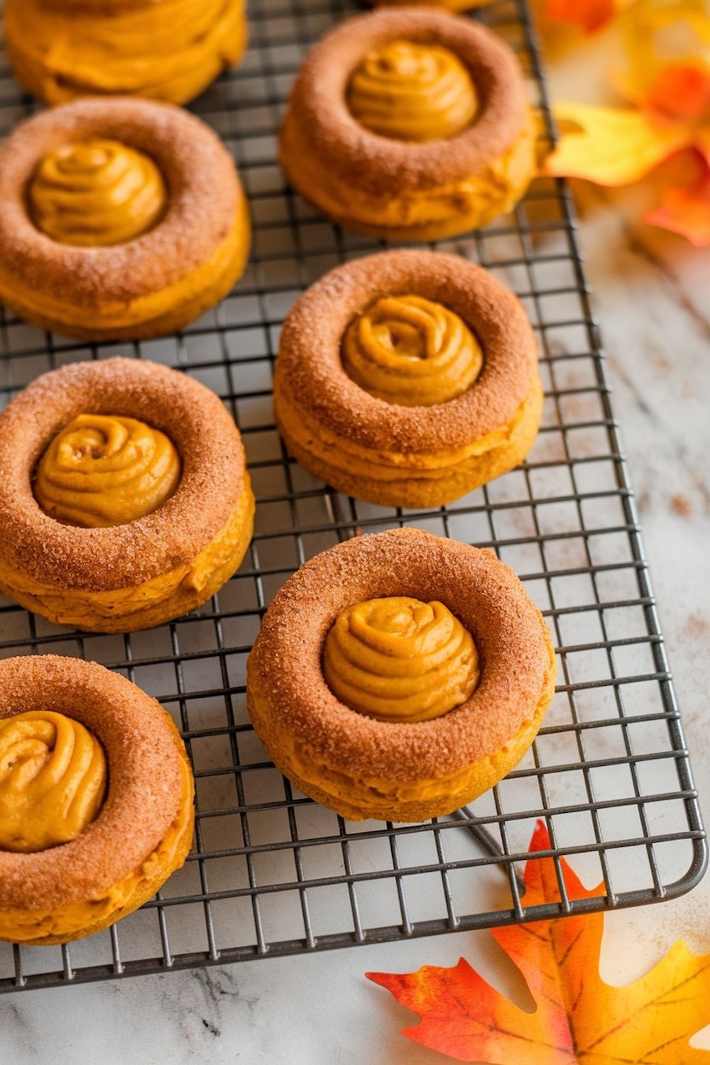 pumpkin spice thumbprint cookies with creamy pumpkin filling arranged on a cooling rack. The cookies are topped with a swirl of pumpkin filling and dusted with cinnamon sugar. In the background, orange autumn leaves are visible, adding a fall theme to the image