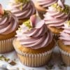 An elegant display of cupcakes with pale pink rose-flavored frosting topped with delicate rose petals and chopped pistachios, arranged on a white porcelain plate with golden accents.