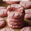 A stack of pink strawberry sugar cookies with a bite taken out of the top cookie, arranged on a brown parchment paper background, showcasing a close-up view of the soft texture of the cookie.