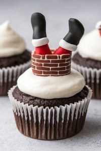 Santa Stuck in the Chimney Dark Chocolate Cupcakes – 24 Days of Christmas Desserts: Playful dark chocolate cupcakes featuring a whimsical Santa decoration, with fondant legs sticking out of a fondant chimney. The fluffy vanilla frosting resembles snow, making these cupcakes a fun and festive treat, straight from "24 Days of Christmas Desserts."