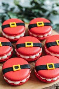 Santa Belt Christmas Macarons – 24 Days of Christmas Desserts: Festive red macarons decorated with a playful Santa belt design, featuring bright yellow buckles and black fondant belts. These fun, bite-sized treats from "24 Days of Christmas Desserts" are filled with creamy vanilla frosting, making them a delightful addition to any holiday dessert spread.