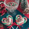 Close-up of festive candy cane heart lollipops, featuring red, white, and green striped candy canes twisted into heart shapes with a creamy white chocolate center, topped with red, green, and white sprinkles. Each lollipop is tied with a red satin ribbon, set against a holiday-themed red background, perfect for Christmas treats or DIY gifts.