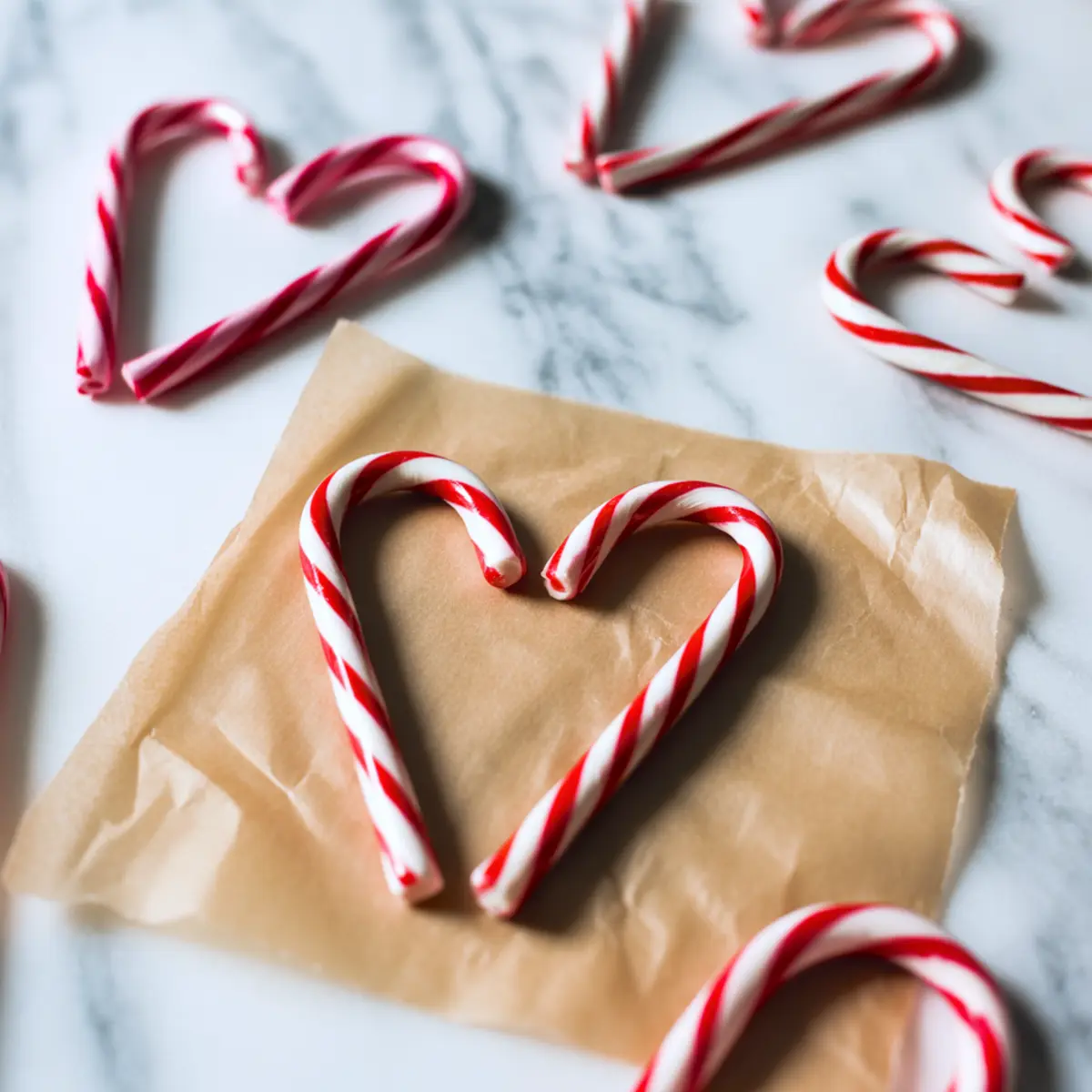 Red and white candy canes arranged into heart shapes on a marble surface, with one heart placed on a piece of parchment paper in preparation for a holiday treat.