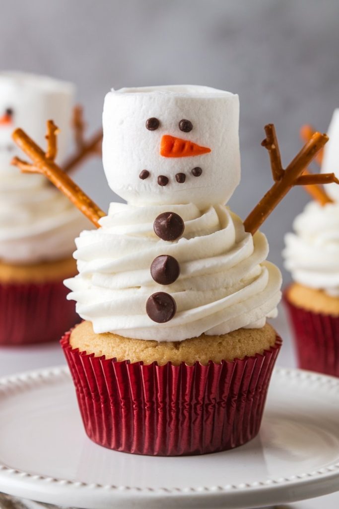 A creatively decorated cupcake designed to look like a snowman, with a marshmallow head, pretzel arms, and chocolate chip features, sitting on a white plate.