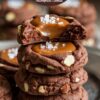 A tall stack of nutty chocolate caramel thumbprint cookies, each topped with gooey caramel and sea salt flakes, set against a vintage backdrop.