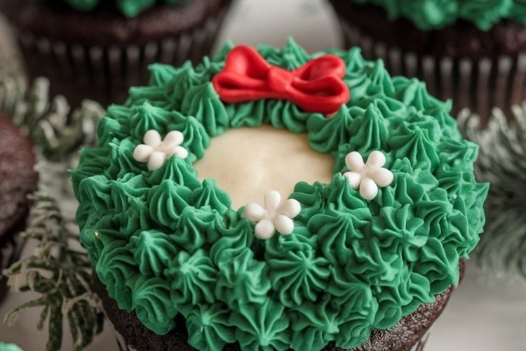 An array of chocolate wreath cupcakes, each topped with a creamy white circle center, green frosting, and red bow, set on a white plate.