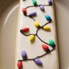 Rectangular sugar cookie decorated with festive Christmas lights. The cookie features a smooth white icing base with piped black lines resembling string lights, adorned with colorful bulb-shaped decorations in red, green, yellow, blue, and purple. This whimsical design captures the holiday spirit, making it a fun and creative addition to Christmas cookie platters.