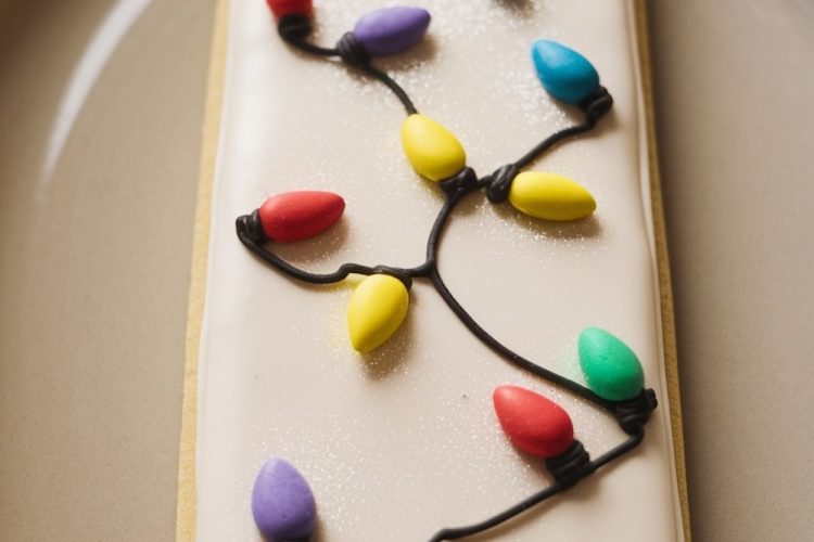 Rectangular sugar cookie decorated with festive Christmas lights. The cookie features a smooth white icing base with piped black lines resembling string lights, adorned with colorful bulb-shaped decorations in red, green, yellow, blue, and purple. This whimsical design captures the holiday spirit, making it a fun and creative addition to Christmas cookie platters.