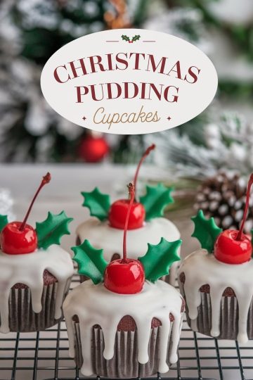 Festive Christmas pudding cupcakes, topped with a smooth white glaze that drips down the sides, adorned with a bright red cherry and green holly leaf decorations. The background features a cozy holiday scene with frosted decorations, enhancing the seasonal theme.