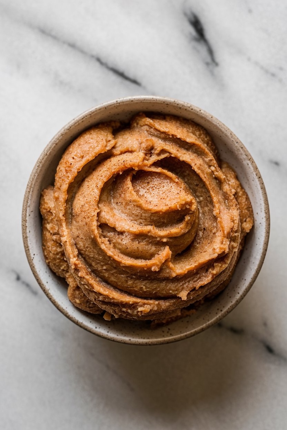 A swirl of cinnamon butter in a small bowl on a white marble surface. The rich, smooth texture of the butter, combined with the warm spices, suggests it's a perfect spread for breakfast pastries or breads, adding a touch of fall-inspired sweetness.