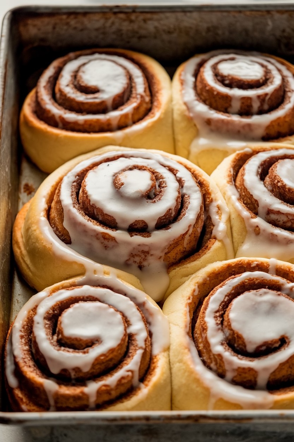 A tray of freshly baked cinnamon rolls topped with a smooth vanilla glaze. The rolls are perfectly swirled, showcasing layers of cinnamon and sugar, with the icing melting into the warm dough. Their golden edges and fluffy texture make them look incredibly soft and inviting, perfect for a sweet breakfast or dessert.