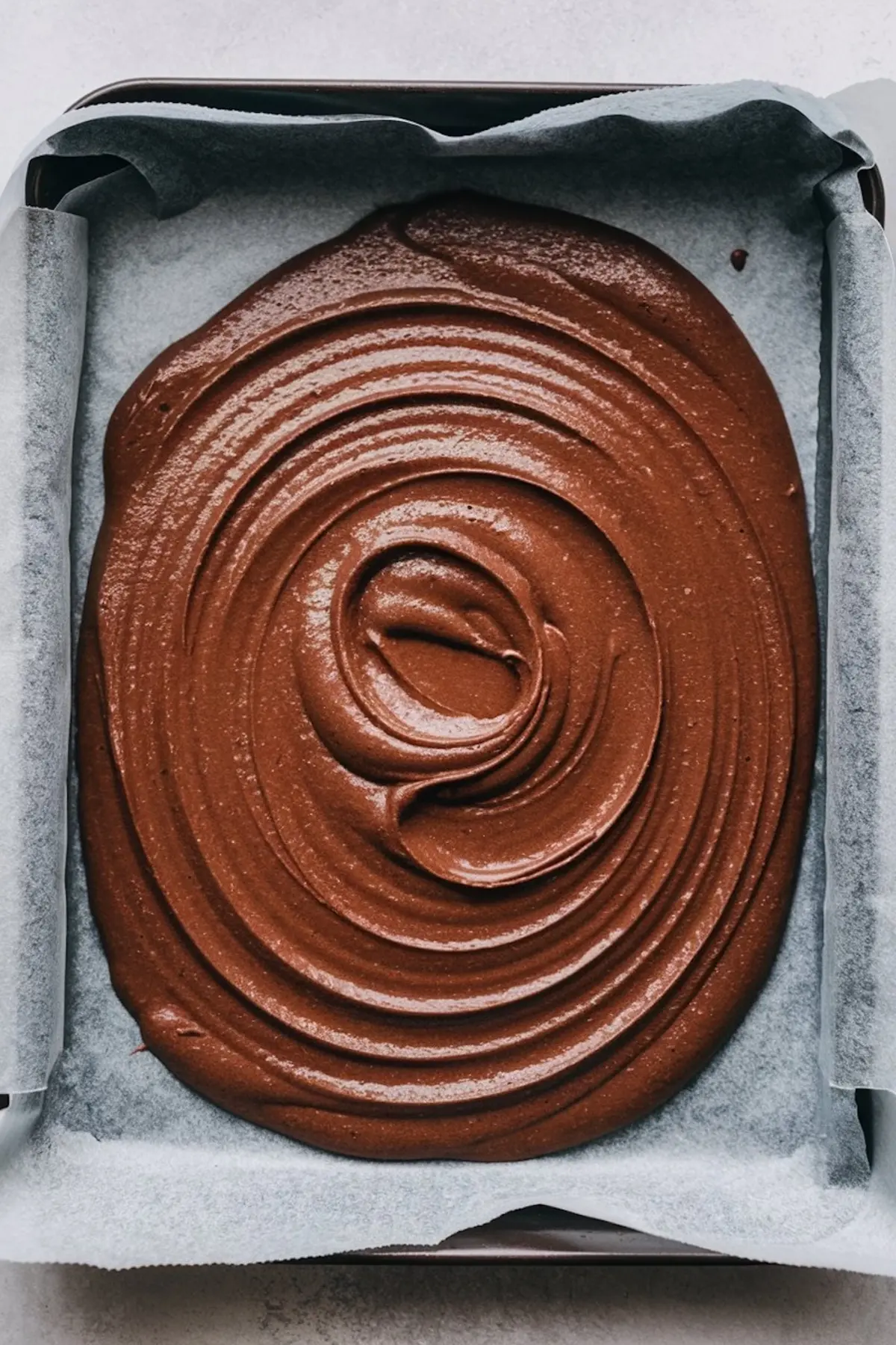 Unbaked chocolate sponge cake batter swirled into a spiral pattern in a parchment-lined baking pan, ready for baking a festive Yule log cake or Buche De Noel.