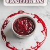 A festive presentation of "Homemade Christmas Cranberry Jam" with a rustic, frosted holiday background. The jam is showcased in a bowl with a decorative cranberry ring around it, emphasizing the rich, deep red color and smooth texture of the jam, perfect for seasonal celebrations.