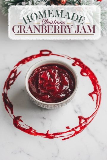 A festive presentation of "Homemade Christmas Cranberry Jam" with a rustic, frosted holiday background. The jam is showcased in a bowl with a decorative cranberry ring around it, emphasizing the rich, deep red color and smooth texture of the jam, perfect for seasonal celebrations.