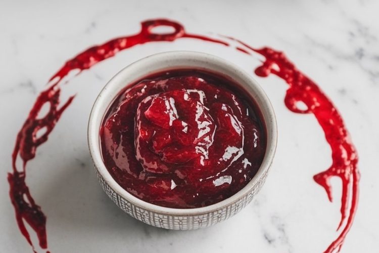 A festive presentation of "Homemade Christmas Cranberry Jam" with a rustic, frosted holiday background. The jam is showcased in a bowl with a decorative cranberry ring around it, emphasizing the rich, deep red color and smooth texture of the jam, perfect for seasonal celebrations.