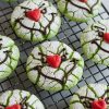 Green crinkle cookies inspired by the Grinch character, perfect for Christmas baking. Cracked, powdered sugar finish that reveals vibrant green underneath, with whimsical chocolate icing faces and bright red heart-shaped noses. Set on a cooling rack, these playful holiday treats are both eye-catching and delicious, ideal for holiday parties, Christmas cookie exchanges.