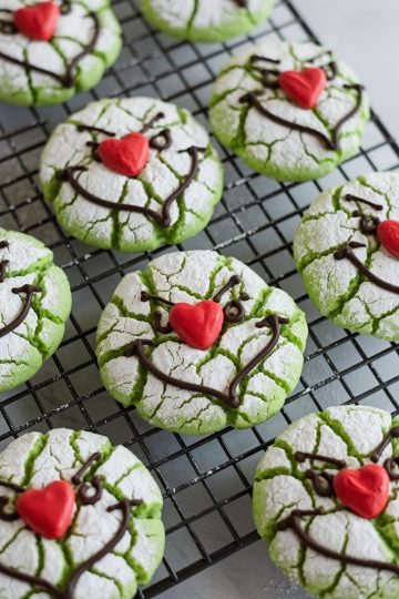 Green crinkle cookies inspired by the Grinch character, perfect for Christmas baking. Cracked, powdered sugar finish that reveals vibrant green underneath, with whimsical chocolate icing faces and bright red heart-shaped noses. Set on a cooling rack, these playful holiday treats are both eye-catching and delicious, ideal for holiday parties, Christmas cookie exchanges.