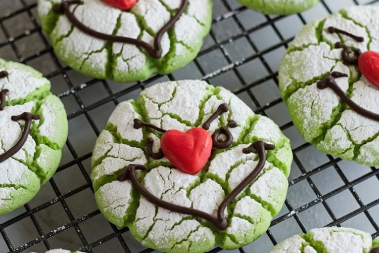 Green crinkle cookies inspired by the Grinch character, perfect for Christmas baking. Cracked, powdered sugar finish that reveals vibrant green underneath, with whimsical chocolate icing faces and bright red heart-shaped noses. Set on a cooling rack, these playful holiday treats are both eye-catching and delicious, ideal for holiday parties, Christmas cookie exchanges.
