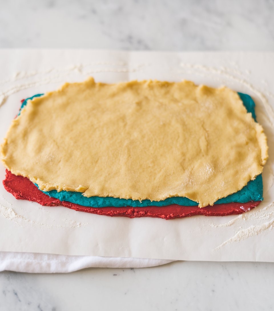 Rolled layers of cookie dough in red, green and natural shades on parchment paper, ready to be shaped into swirls. This behind-the-scenes look shows the preparation process, where the dough is stacked and rolled before being cut into festive pinwheel cookies. Perfect for a step-by-step holiday baking tutorial.