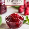 Close-up view of raspberry jam in a clear glass bowl, with fresh raspberries and mint leaves in the background, accompanied by a stylized text overlay 'Homemade Raspberry Jam Recipe.