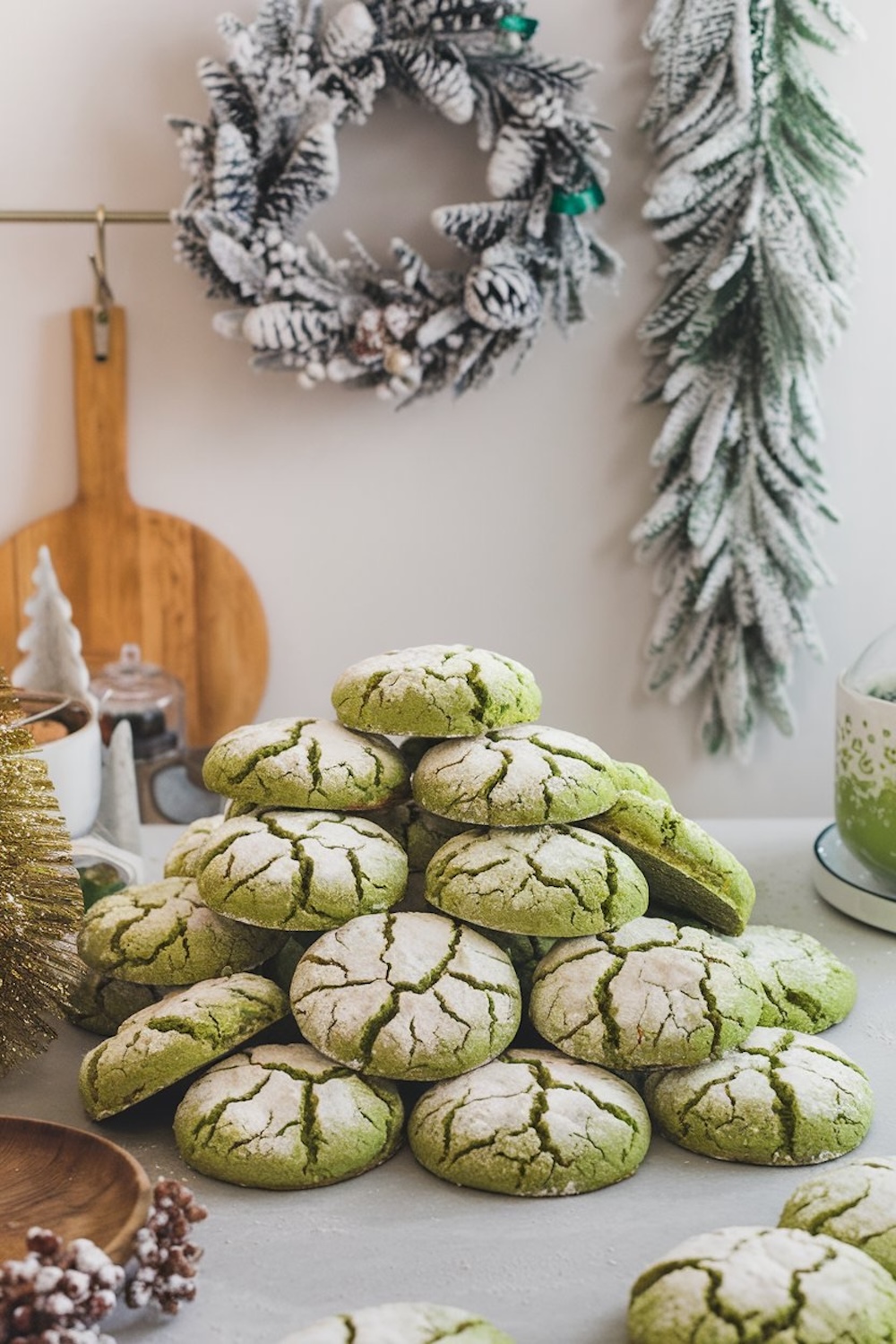 A festive display of matcha crinkle cookies piled on a gray countertop, surrounded by holiday decorations including a snowy wreath and a frosted garland, creating a cozy, seasonal baking scene.