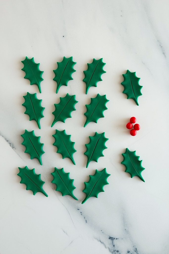 Several green fondant holly leaves placed on a white marble background, accompanied by small red fondant berries, set to decorate no-bake Rice Krispie Christmas Puddings.