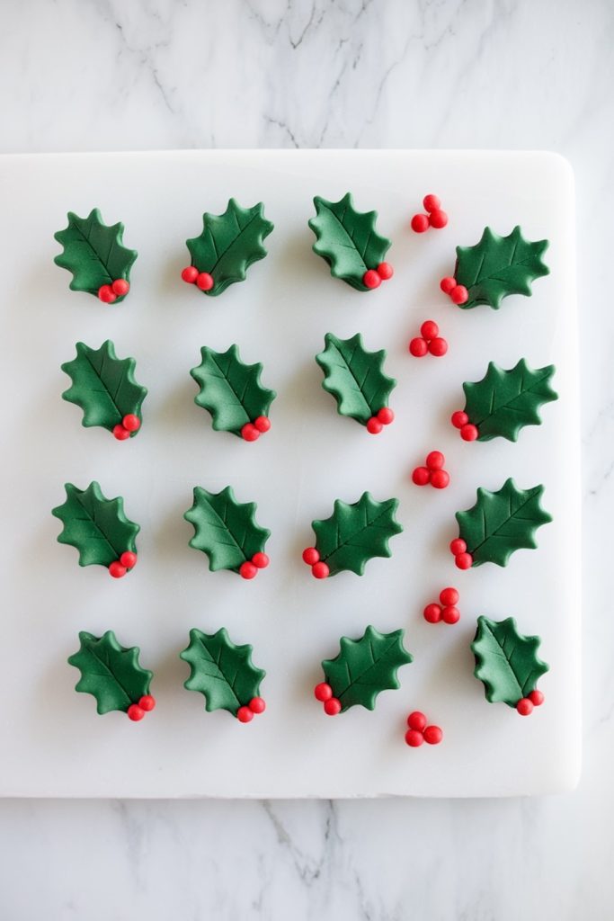 Green holly leaf fondant decorations arranged neatly on a white board, each adorned with bright red berry clusters for festive no-bake Rice Krispie Christmas Puddings.