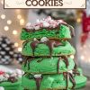 A stack of vibrant green peppermint hot chocolate cookies, with one cookie on top split open to reveal a soft, festive green interior. Each cookie is generously drizzled with melted chocolate and sprinkled with mini marshmallows and crushed peppermint candy, adding a festive, holiday-inspired touch. The background is softly lit with warm holiday lights, adding a cozy, Christmas ambiance to the scene.