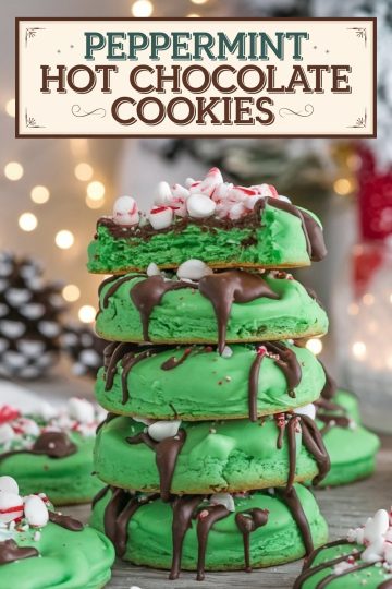 A stack of vibrant green peppermint hot chocolate cookies, with one cookie on top split open to reveal a soft, festive green interior. Each cookie is generously drizzled with melted chocolate and sprinkled with mini marshmallows and crushed peppermint candy, adding a festive, holiday-inspired touch. The background is softly lit with warm holiday lights, adding a cozy, Christmas ambiance to the scene.