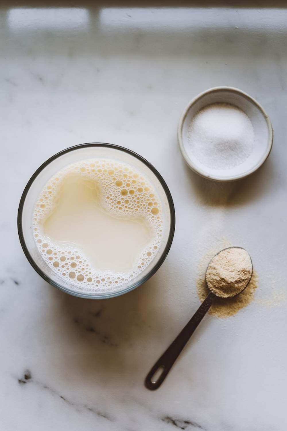A glass of foamy milk next to a small bowl of sugar and a spoonful of yeast on a marble countertop, essential ingredients for dough preparation. The setup captures a calm, minimalist aesthetic, emphasizing the importance of fresh ingredients in baking.