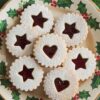 Elegant Linzer cookies on a Christmas plate featuring holly patterns, with powdered sugar and raspberry jam filling star and heart-shaped centers, conveying a festive holiday spirit.
