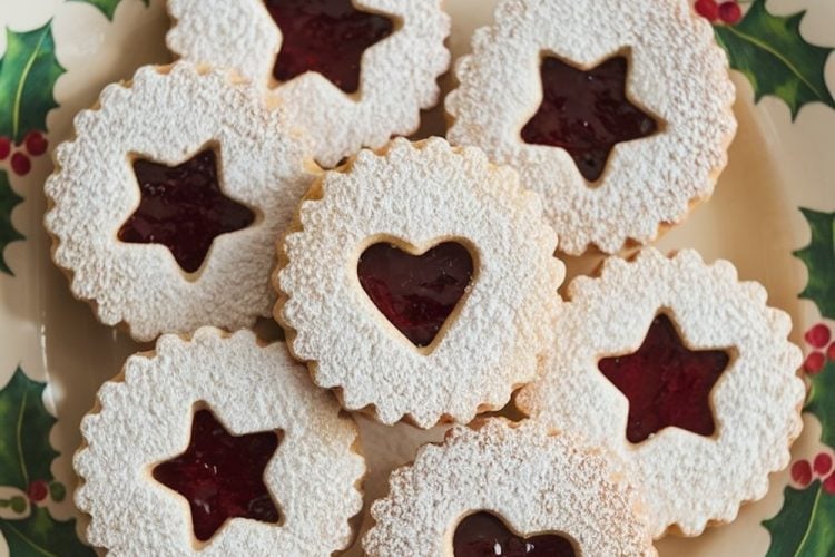Elegant Linzer cookies on a Christmas plate featuring holly patterns, with powdered sugar and raspberry jam filling star and heart-shaped centers, conveying a festive holiday spirit.
