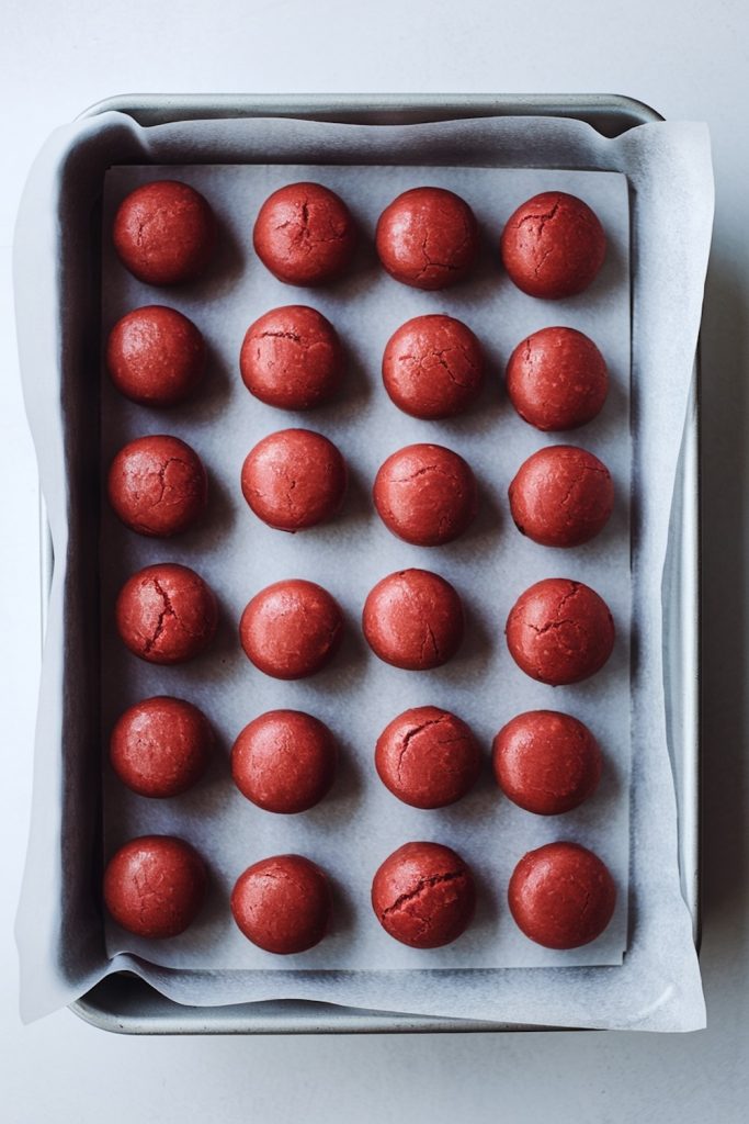 Rows of round red velvet cookie dough balls arranged neatly on a parchment-lined baking tray, ready to be baked.