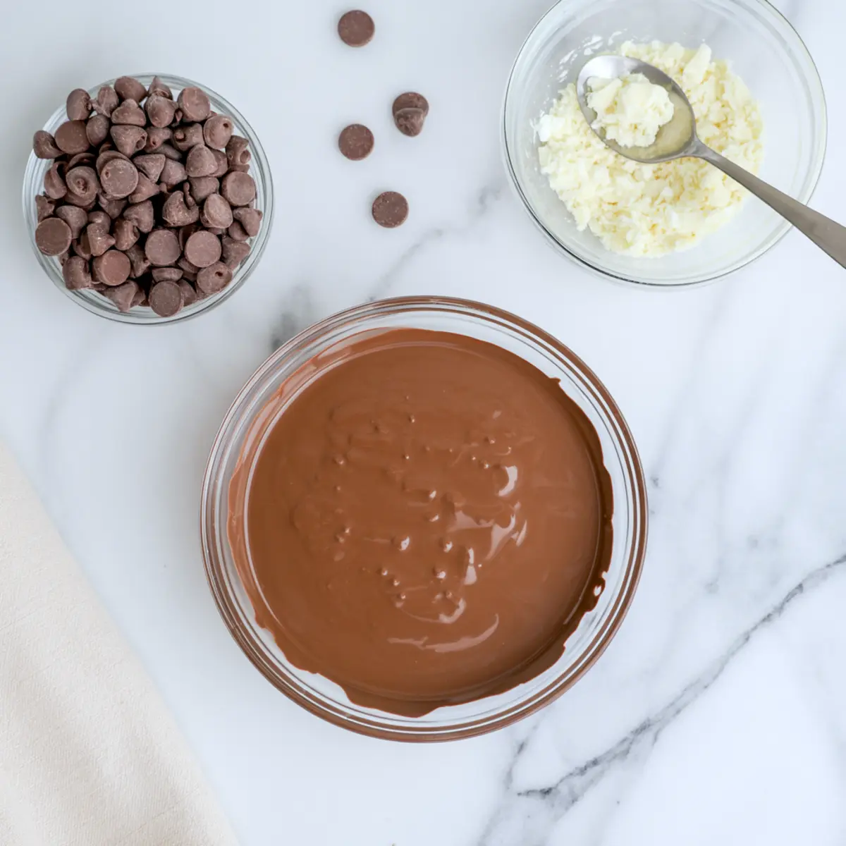 Bowls of milk chocolate chips, chopped white candy coating, and melted chocolate on a white marble background prepared for cookie decorating.