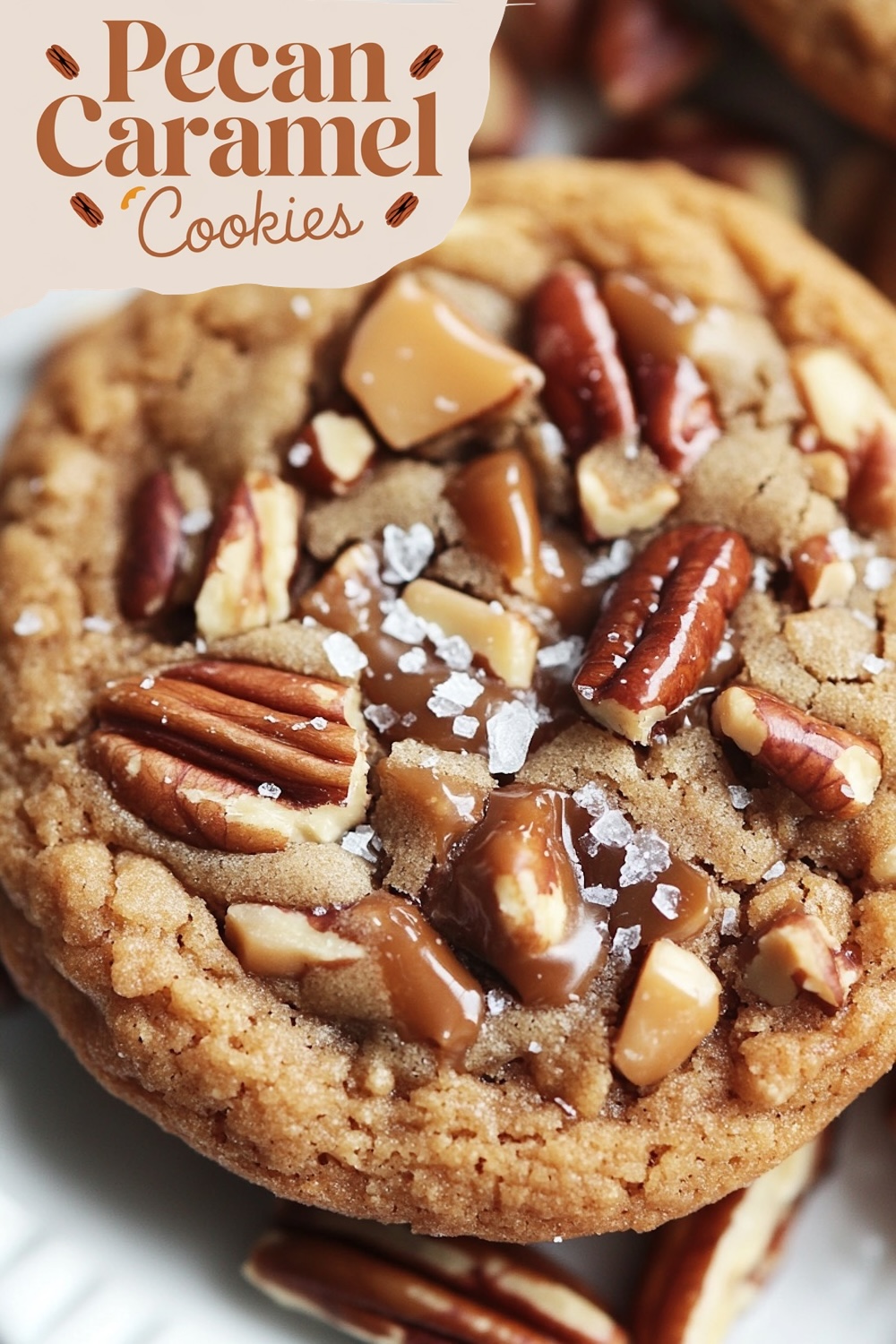 A single pecan caramel cookie up close, showing chunky pieces of pecan and caramel chunks melted on top. The cookie is soft, and the caramel appears gooey, with a dusting of flaky sea salt enhancing the texture.