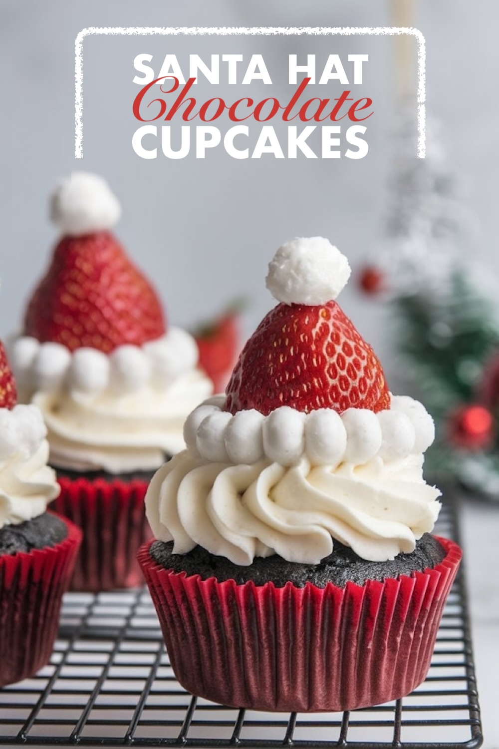 A close-up shot of Christmas-themed cupcakes topped with strawberries as Santa hats, surrounded by glowing lights and holiday decor. The fluffy vanilla frosting makes the design pop.