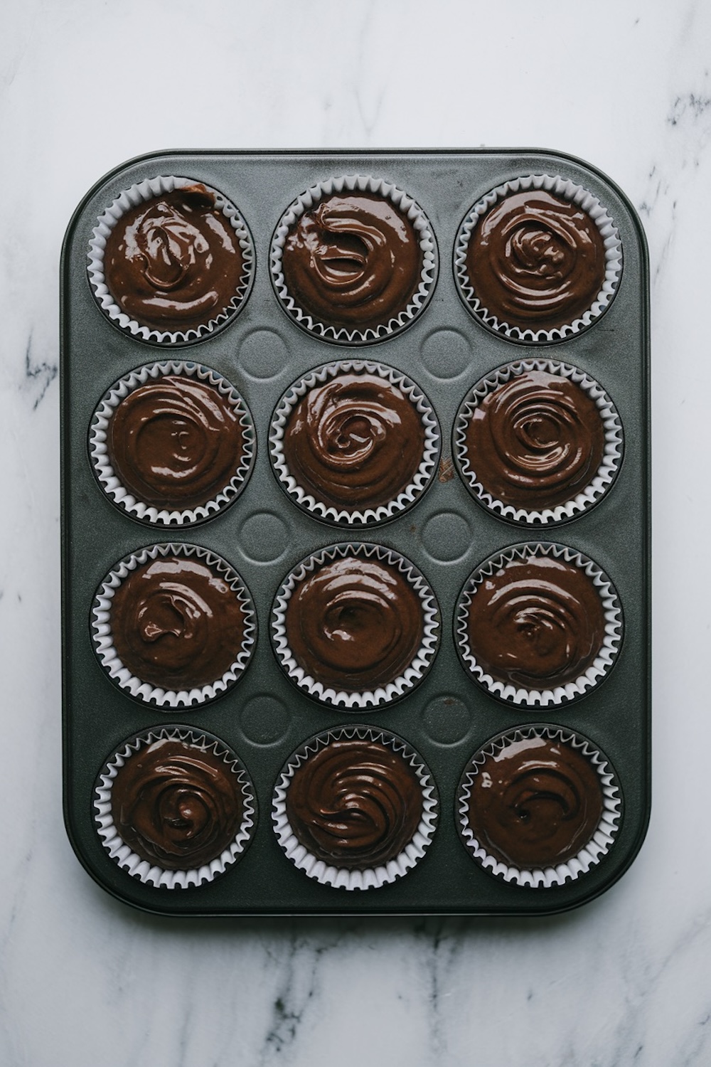 Cupcake batter in a dark chocolate shade is poured into a 12-cup muffin tin lined with paper wrappers. The batter has a glossy, rich texture, perfect for baking moist cupcakes with a decadent chocolate flavor.