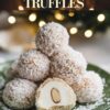 A green plate stacked with white coconut-covered almond truffles. The truffles are dusted with coconut flakes, and one truffle is sliced open, showing the almond center inside a white creamy filling. Christmas tree lights blur softly in the background.