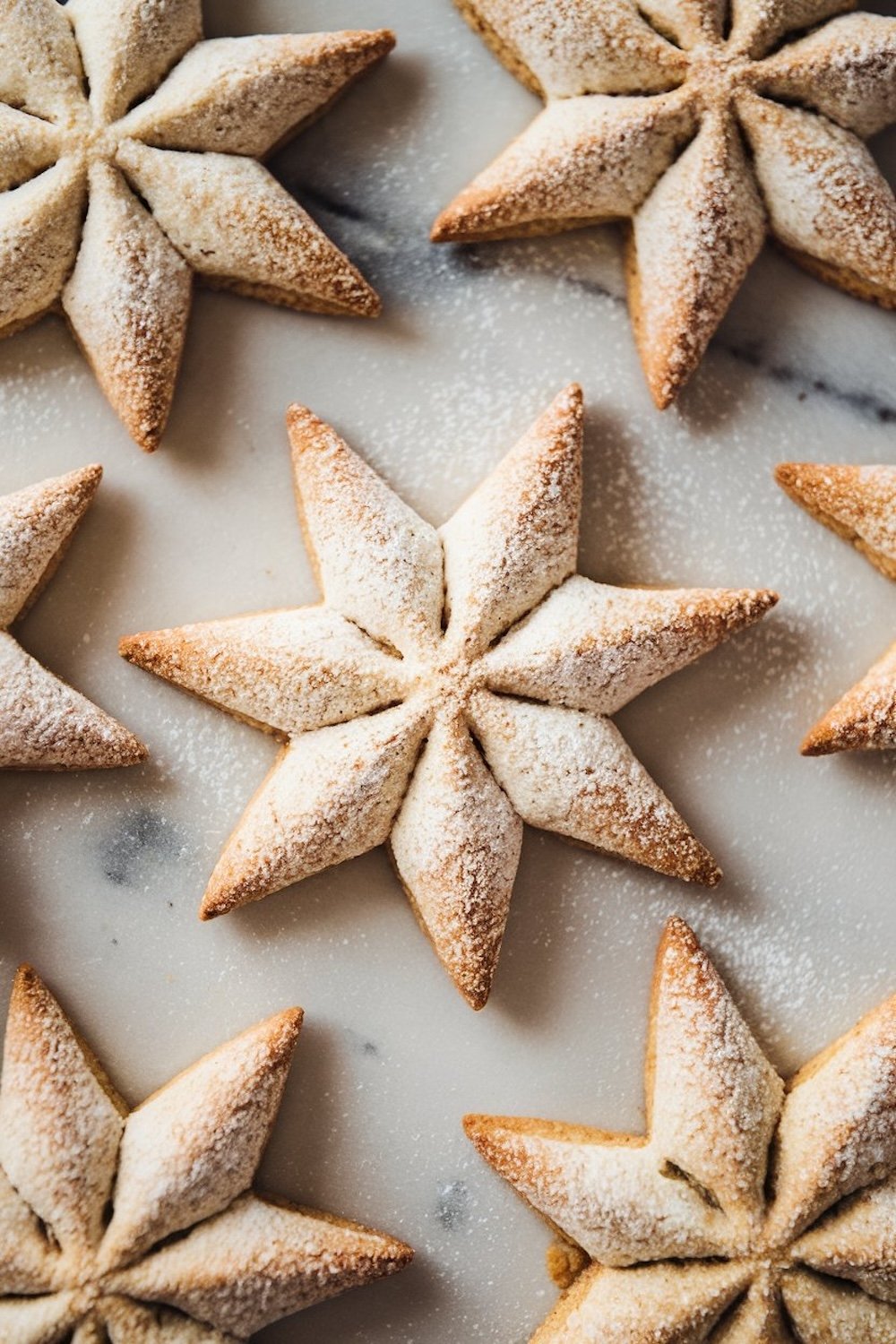 Golden-brown snowflake-shaped cookies dusted with powdered sugar, arranged on a marble surface. Each cookie features detailed, crisp points and a soft, airy texture, evoking a festive, winter wonderland aesthetic. The light dusting of sugar adds a snowy effect, enhancing the holiday appeal of these Sparkling Snowflake Christmas Cookies. Perfect for holiday gatherings, these cookies bring a touch of elegance and seasonal charm to any dessert table.