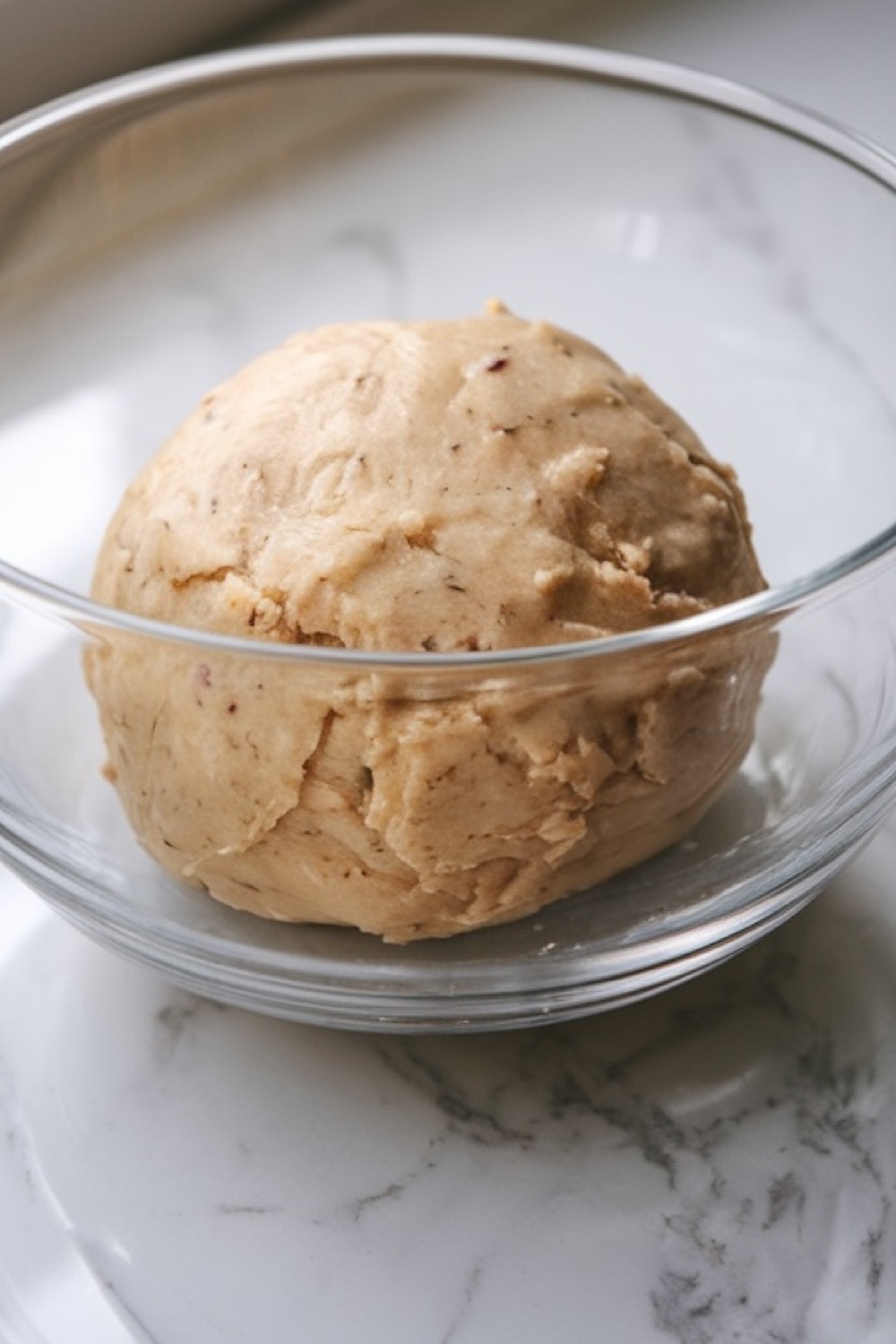 A glass bowl containing a ball of freshly mixed cookie dough for Sparkling Snowflake Christmas Cookies. The dough has a smooth, pliable texture, ready to be rolled out and cut into festive shapes. The soft, light color of the dough complements the clean, marble background, hinting at the buttery richness and delicate flavor that will make these cookies perfect for holiday gatherings.